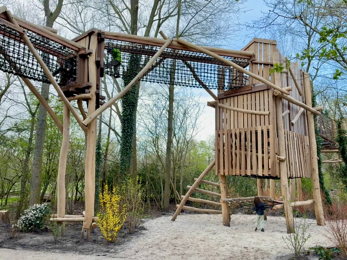 Kids playing on large wooden tower structure with elevated rope and net walkway through the trees