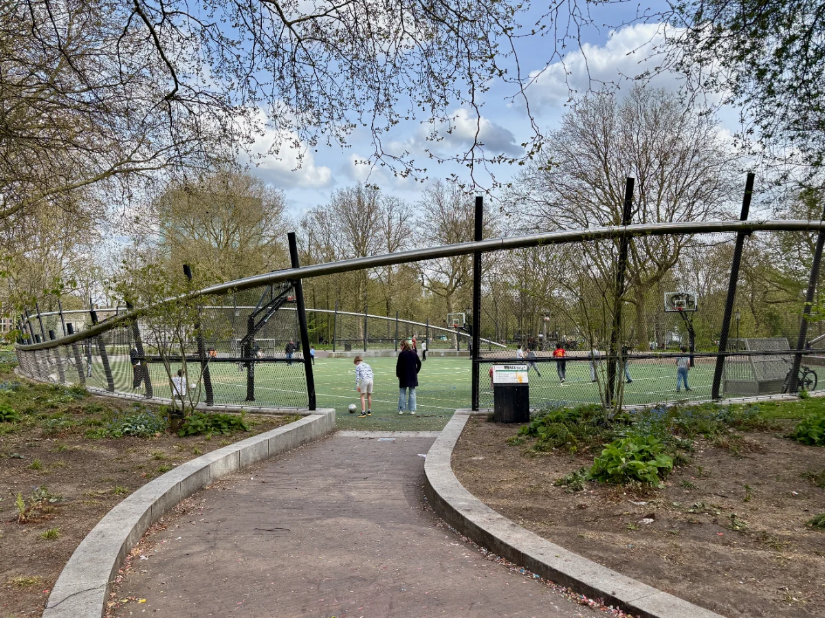 Kids and adults play on green football pitch in park setting with curvy enclosure and trees surrounding