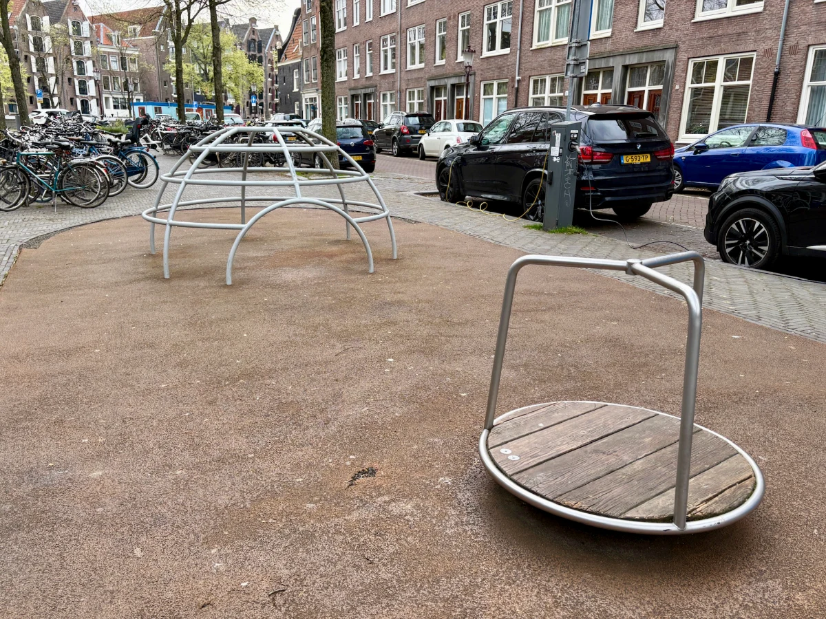 Metal and wood merry-go-round and metal climbing igloo with cars and buildings behind