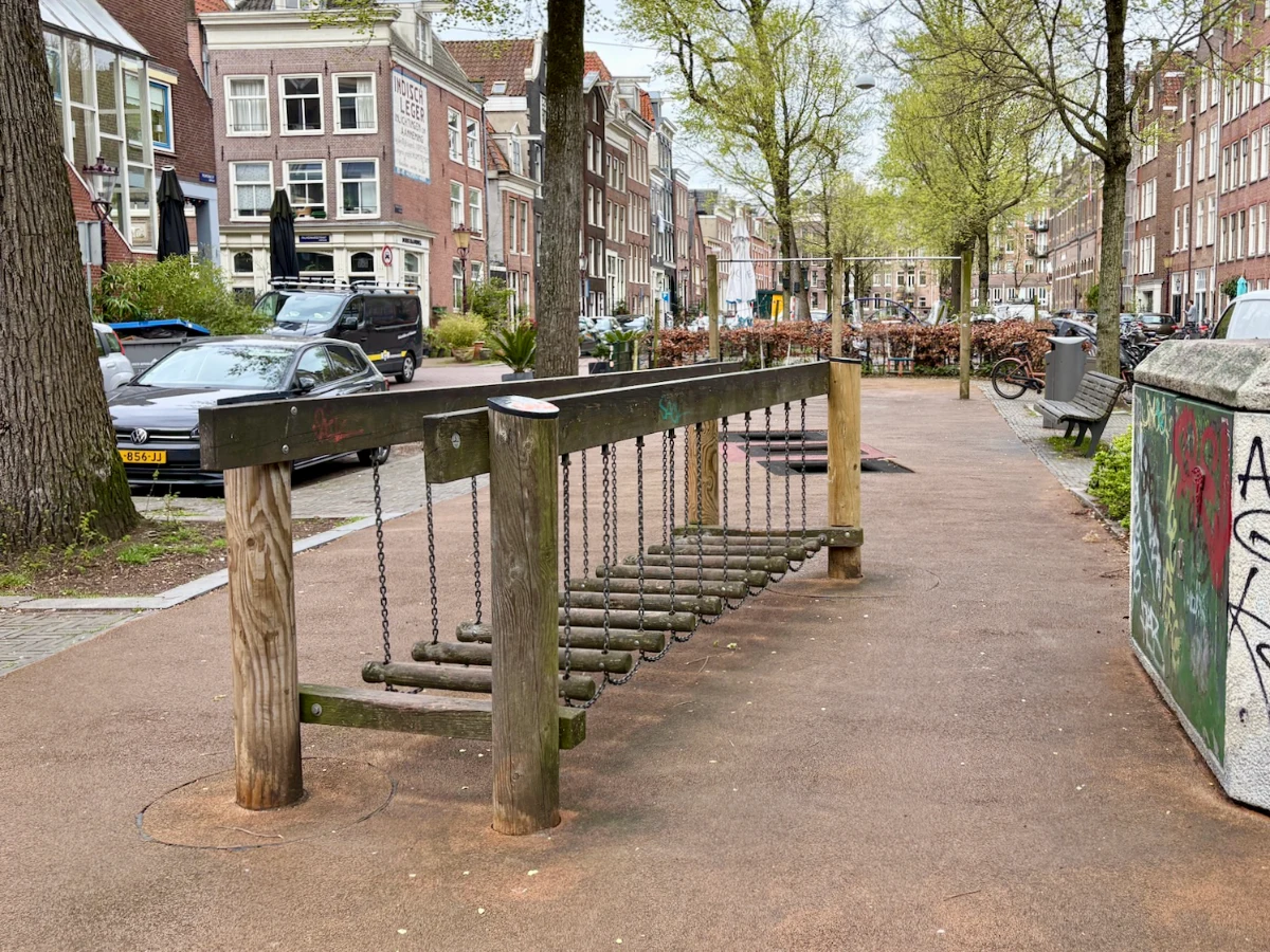 Wooden balance bridge in small playground surrounded by mature trees and parked cars