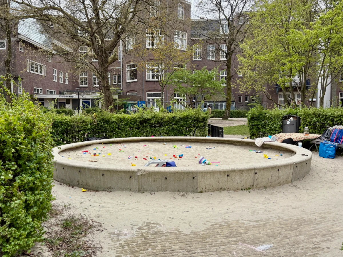 Round sand box with raised concrete sides and lots of sand toys, surrounded by hedges in a residential area