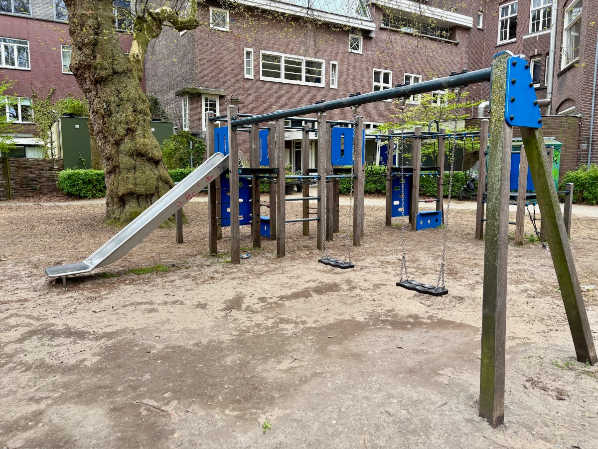 Wooden and blue play structure with swings, slide, and rope nets on sand and next to a large mature tree