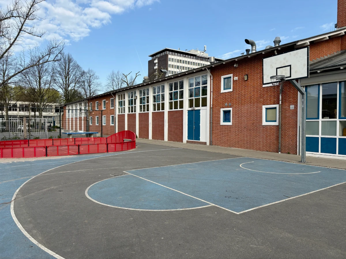 Blue and black paved basketball court next to a brick building with small enclosed football pitch adjacent