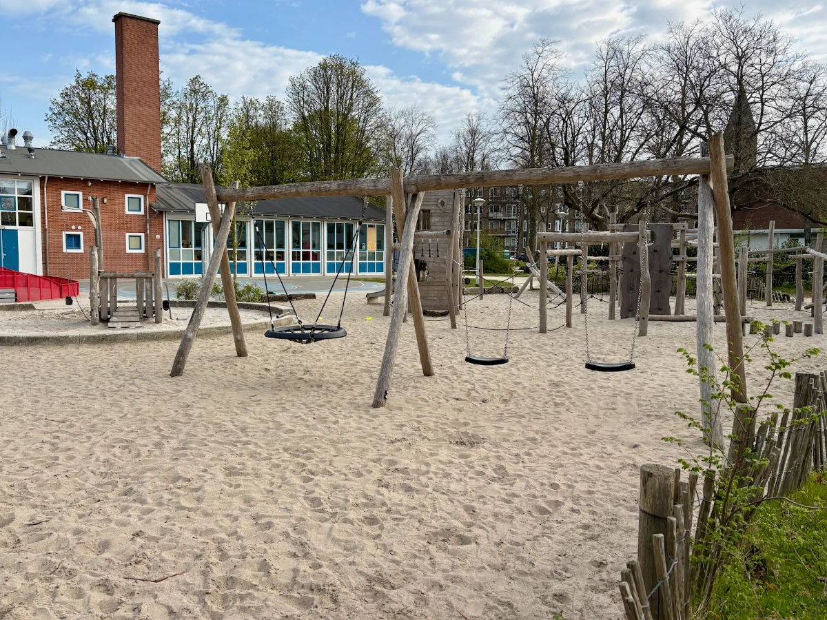 Sandy playground with swings and nest swing and red brick and blue building behind