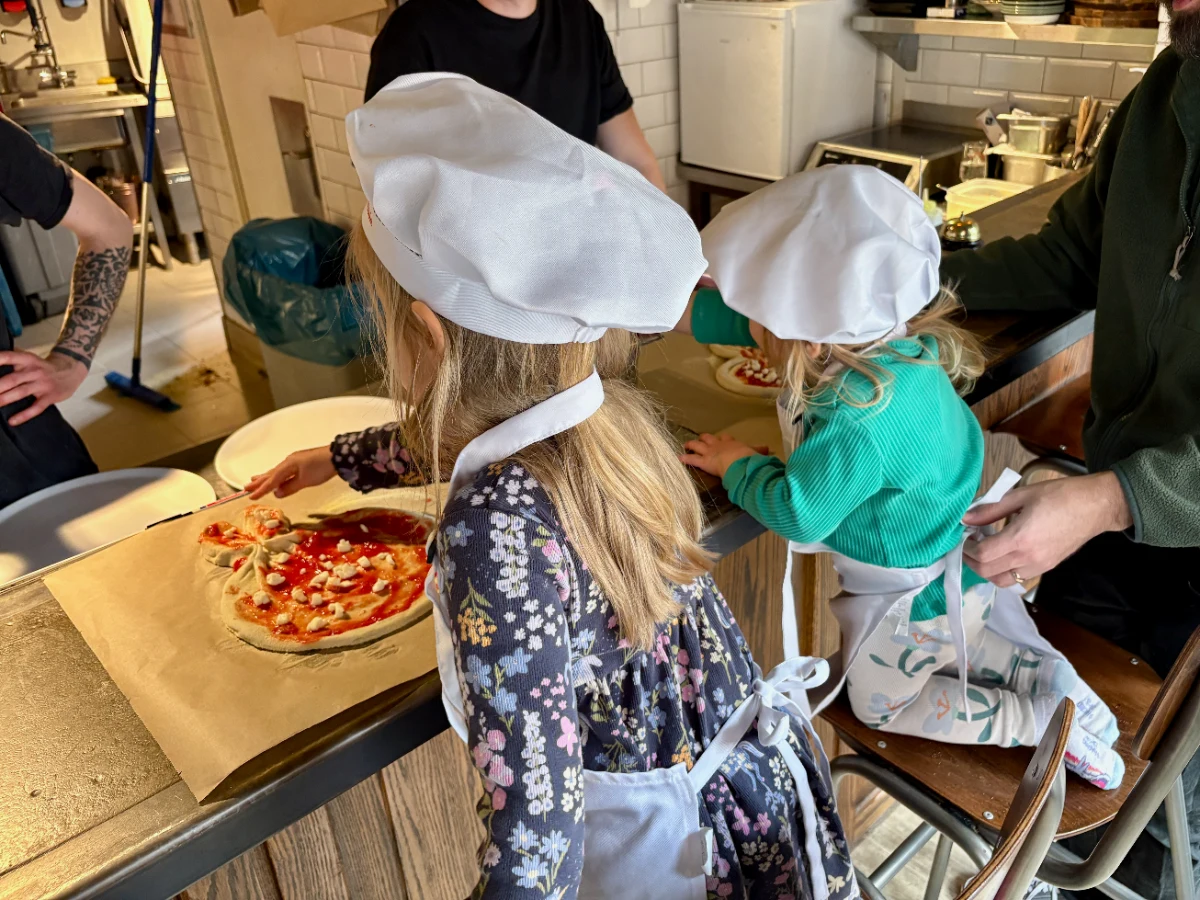 Two kids in chef hats and aprons making pizzas at a counter