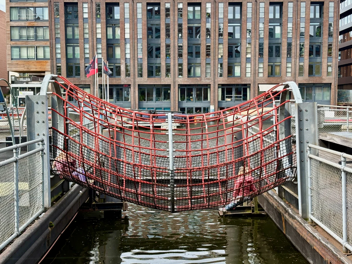 Two kids climbing across a red rope bridge connecting two barges with water underneath