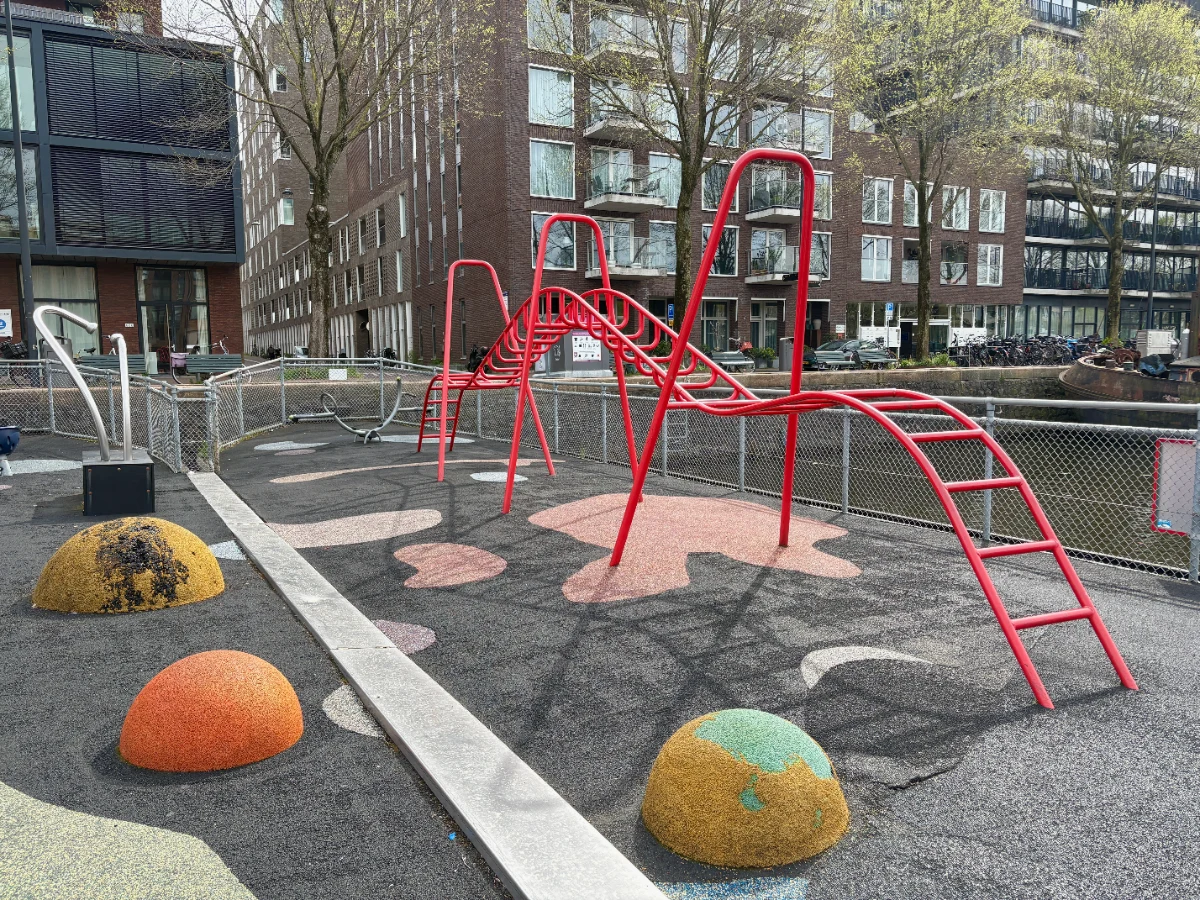 Modern red undulating monkey bar play structure with metal rocker and residential buildings around