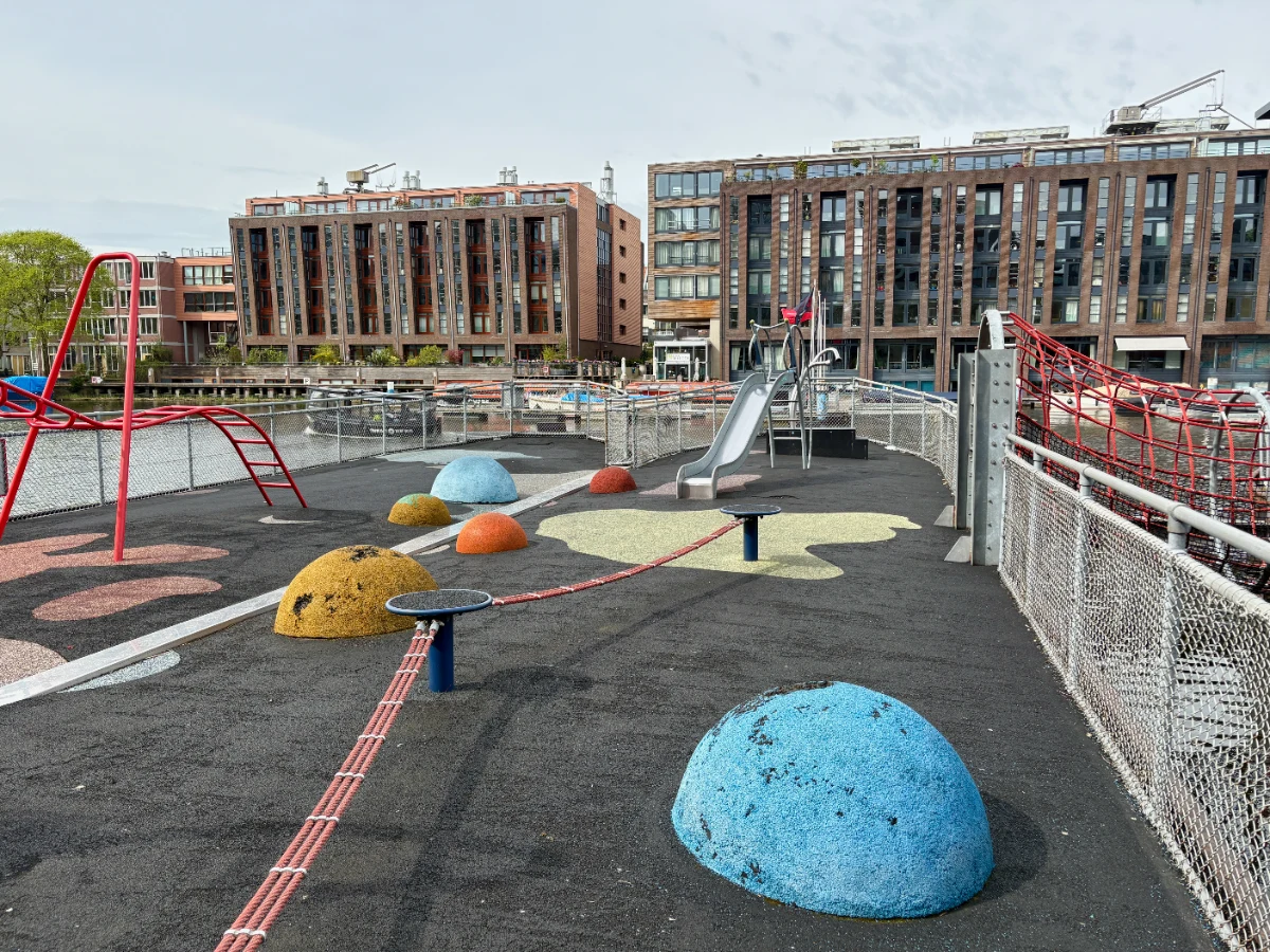 Barge playground on the water with red ladder, balance rope, slide, and blue, yellow, and orange sheres