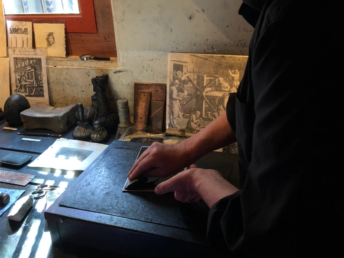 Man applying ink to a copper plate with etchings and various artist tools on the table