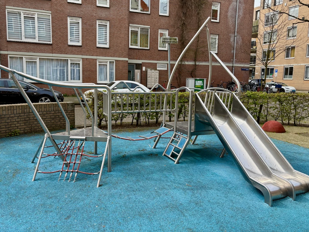 Metal play structure for little kids with rope climbing nets, bridge, and twin slides set on blue rubberized surface next to residential building