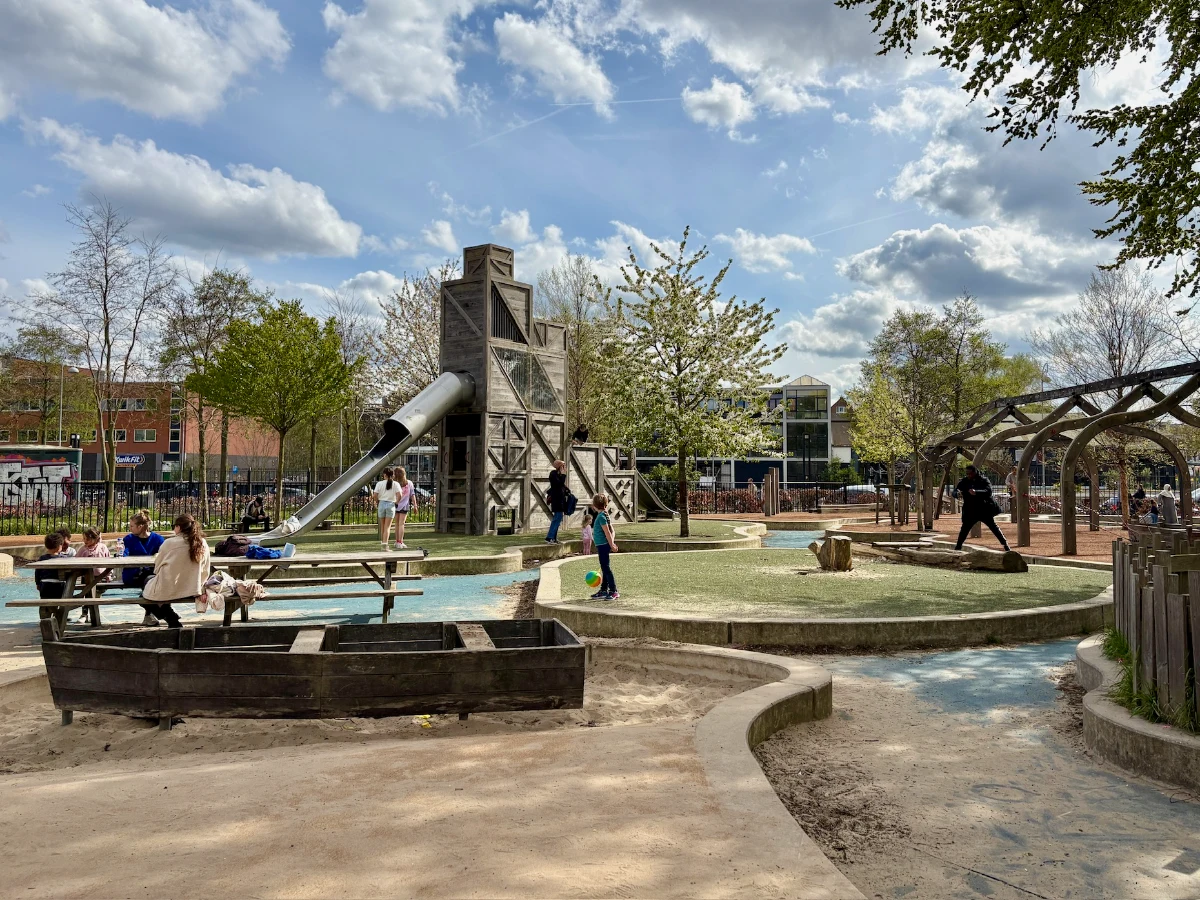 Kids and parents playing on playground with tall wooden tower with slide, wooden boat, and raised astroturf play zones with blue pathways