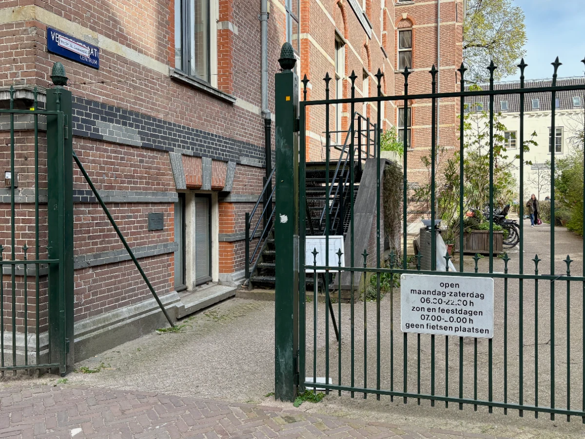 Wrought iron gate leading to courtyard with large brick building and sign posted outside with opening hours