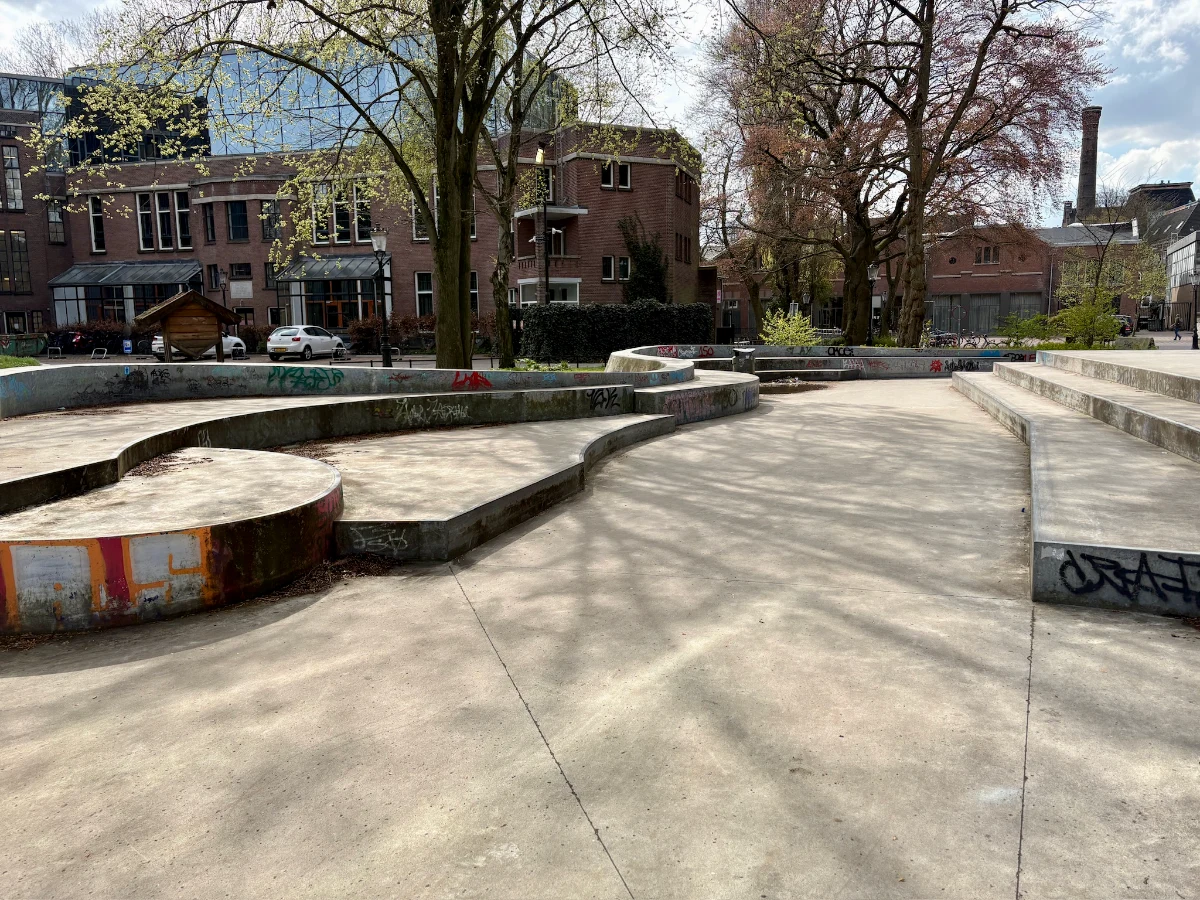 Shaded skatepark with multiple levels and stairs within buildings and trees behind
