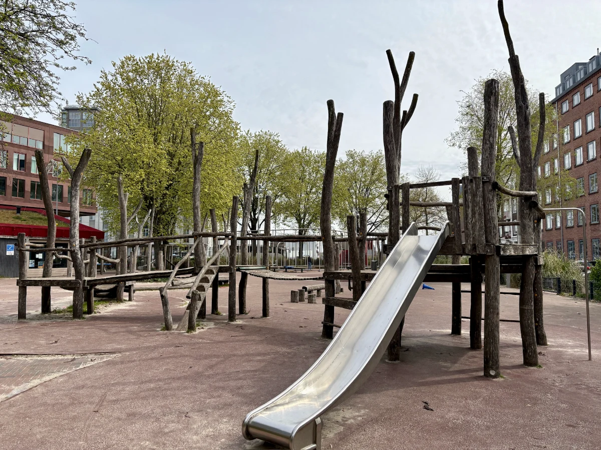 Natural wooden playground structure with bridge, ladder, and slide and large trees behind