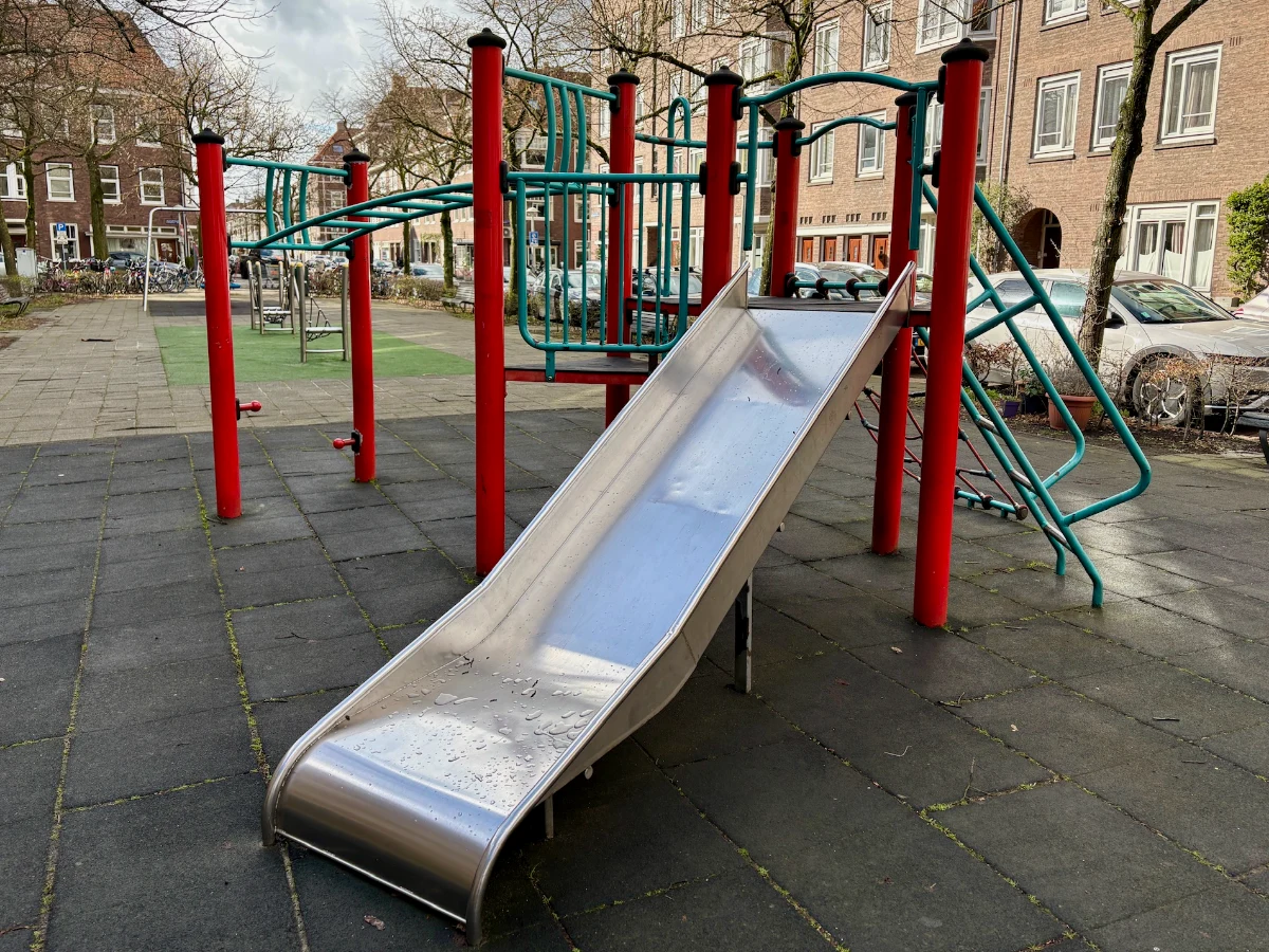 Red and green metal climbing structure with slide and residential buildings behind