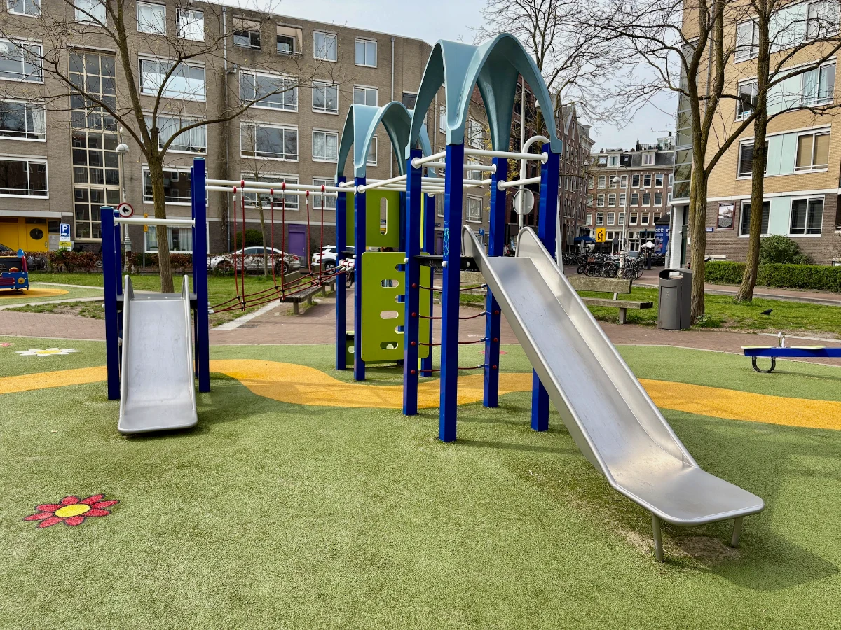 Blue and green playground structure with two slides and a rope bridge set on a small residential square